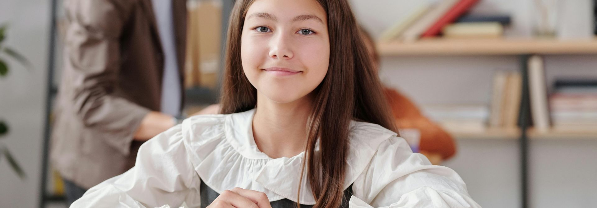 A young student smiling at her desk in a classroom with books and materials around her.